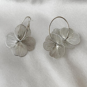 K&ouml;nnte beinhalten: Ein Paar silberfarbene Blumenohrringe. Jeder Ohrring hat ein dreidimensionales Blumendesign mit filigranen, linearen Details. Die Ohrringe befinden sich auf einem wei&szlig;en Hintergrund.