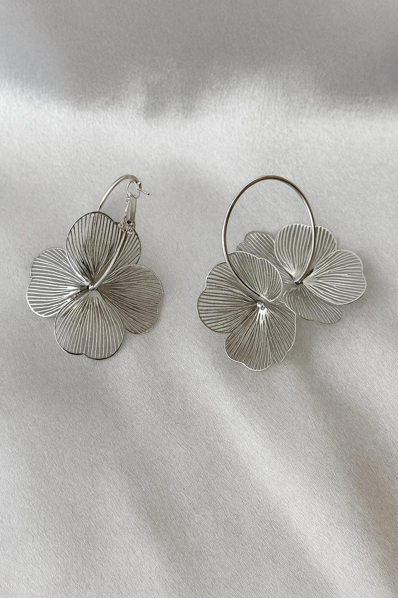 K&ouml;nnte beinhalten: Ein Paar silberfarbene Blumenohrringe. Jeder Ohrring hat ein dreidimensionales Blumendesign mit filigranen, linearen Details. Die Ohrringe befinden sich auf einem wei&szlig;en Hintergrund.