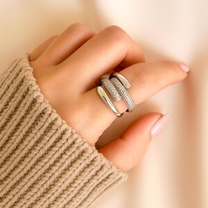 May include: A silver-toned ring with a unique, layered design. One band is encrusted with small, sparkling stones, while the other bands are smooth. The ring is worn on a finger, showcasing its modern and elegant style.