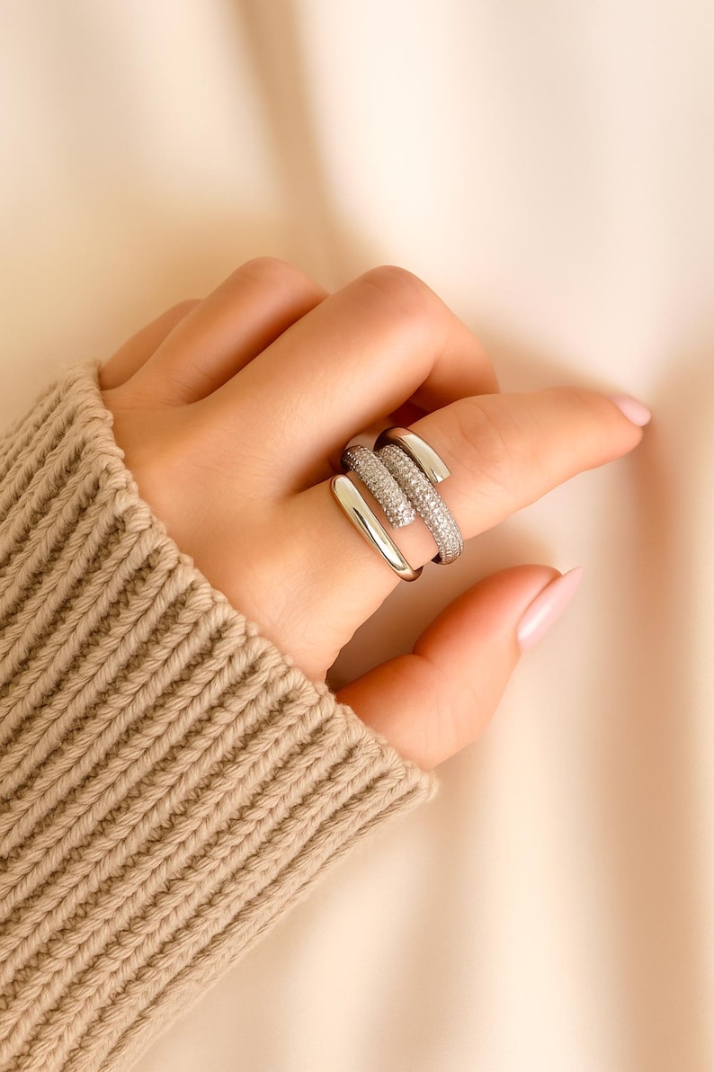 May include: A silver-toned ring with a unique, layered design. One band is encrusted with small, sparkling stones, while the other bands are smooth. The ring is worn on a finger, showcasing its modern and elegant style.
