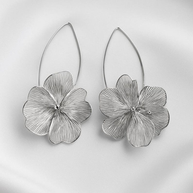 K&ouml;nnte beinhalten: Ein Paar silberfarbene Blumenohrringe. Jeder Ohrring hat ein detailliertes, durchbrochenes Blumendesign, das an einem tropfenf&ouml;rmigen Draht h&auml;ngt. Die Blumen haben ein strukturiertes, geripptes Aussehen mit einer kleinen Perle in der Mitte.