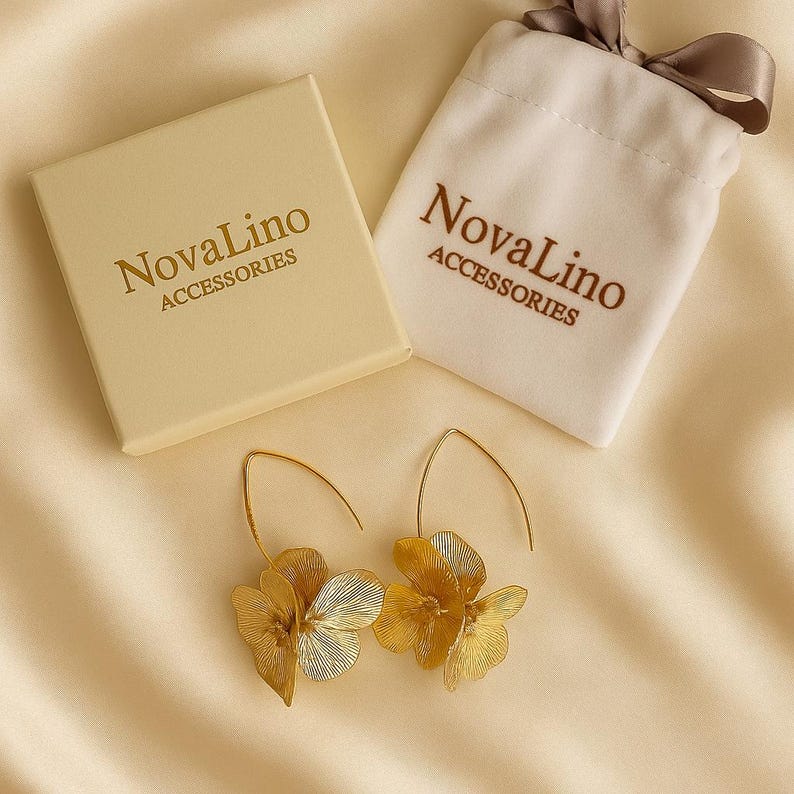 K&ouml;nnte beinhalten: Goldfarbene Blumenohrringe mit einer passenden cremefarbenen Schachtel und einem Beutel. Auf der Schachtel und dem Beutel steht "NovaLino ACCESSORIES". Die Ohrringe haben ein zartes, geschichtetes Bl&uuml;tenblattdesign.