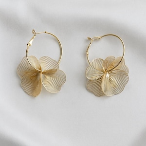 K&ouml;nnte beinhalten: Ein Paar goldfarbene Creolen, jeweils mit einem filigranen, mehrschichtigen Blumendesign. Die Bl&uuml;ten bestehen aus d&uuml;nnen, parallelen Linien, die einen strukturierten, durchbrochenen Effekt erzeugen. Die Ohrringe werden auf wei&szlig;em Hintergrund pr&auml;sentiert.