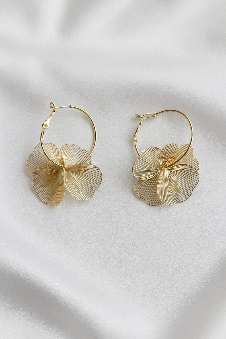 K&ouml;nnte beinhalten: Ein Paar goldfarbene Creolen, jeweils mit einem filigranen, mehrschichtigen Blumendesign. Die Bl&uuml;ten bestehen aus d&uuml;nnen, parallelen Linien, die einen strukturierten, durchbrochenen Effekt erzeugen. Die Ohrringe werden auf wei&szlig;em Hintergrund pr&auml;sentiert.