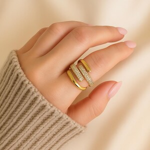 May include: A gold-colored ring with a unique, layered design. One band is encrusted with small, sparkling stones, while the other bands are smooth. The ring is worn on a finger, showcasing its elegant and modern style.