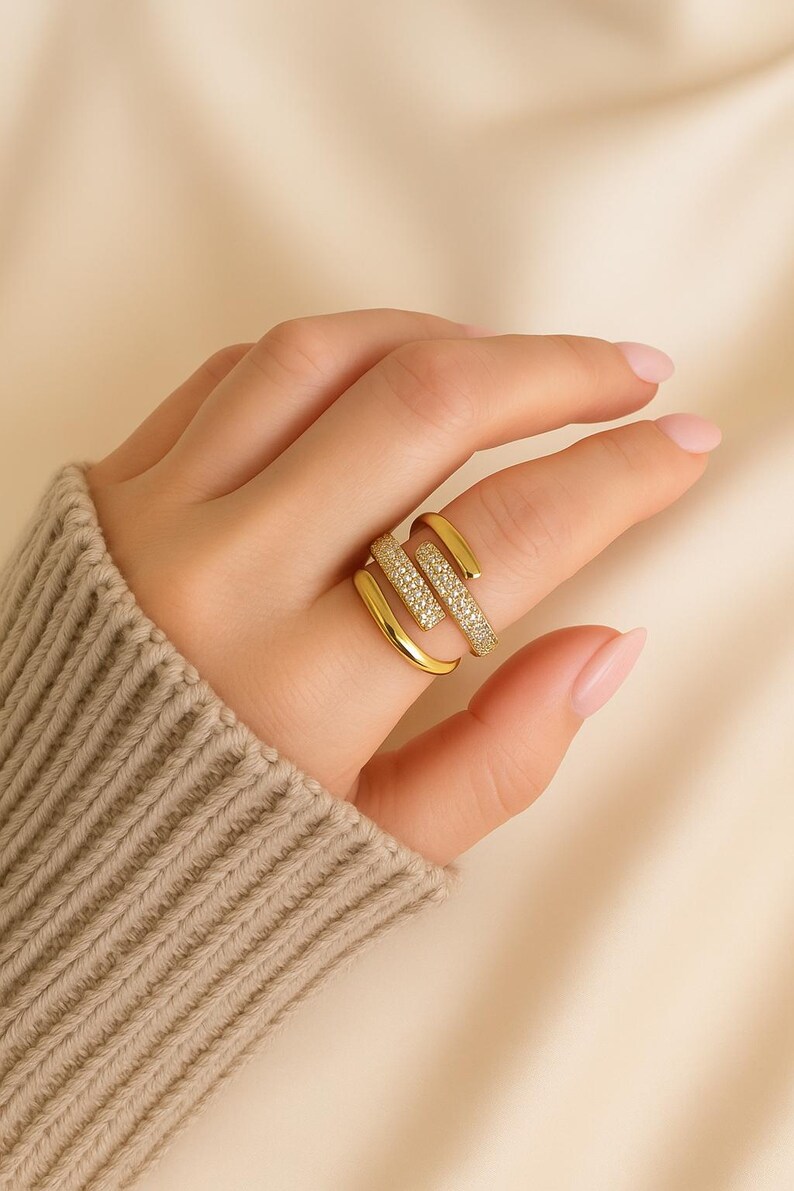 May include: A gold-colored ring with a unique, layered design. One band is encrusted with small, sparkling stones, while the other bands are smooth. The ring is worn on a finger, showcasing its elegant and modern style.
