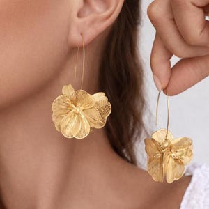 May include: Gold-colored floral earrings. The earrings feature a delicate, layered flower design with intricate detailing. The earrings are suspended from a thin, curved wire. The earrings are shown against a blurred background.