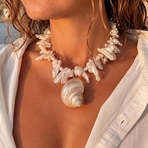 May include: A necklace featuring a large, spiraled, iridescent seashell pendant suspended from a strand of irregular, white, lustrous shell fragments and small round pearls. The necklace rests against the neck, showcasing a natural, pearlescent finish.