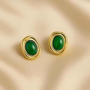 May include: A pair of gold-toned stud earrings with oval, emerald-green centers. The earrings have a smooth, polished finish and a slightly irregular, organic shape. The background is a neutral, cream-colored fabric.
