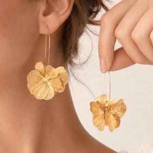 K&ouml;nnte beinhalten: Goldfarbene Blumenohrringe. Die Ohrringe haben ein geschichtetes, dreidimensionales Blumendesign mit detaillierten Bl&uuml;tenblattstrukturen. Sie sind an d&uuml;nnen, gebogenen Ohrhaken aufgeh&auml;ngt, was einen zarten und eleganten Look erzeugt. Die Ohrringe werden an einem Ohr und einer Hand gezeigt.