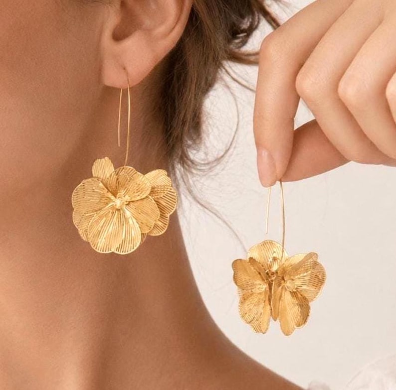 K&ouml;nnte beinhalten: Goldfarbene Blumenohrringe. Die Ohrringe haben ein geschichtetes, dreidimensionales Blumendesign mit detaillierten Bl&uuml;tenblattstrukturen. Sie sind an d&uuml;nnen, gebogenen Ohrhaken aufgeh&auml;ngt, was einen zarten und eleganten Look erzeugt. Die Ohrringe werden an einem Ohr und einer Hand gezeigt.