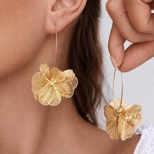 K&ouml;nnte beinhalten: Goldfarbene Blumenohrringe. Die Ohrringe haben ein filigranes, durchbrochenes Design in Form einer Blume. Sie sind an einem d&uuml;nnen, gebogenen Draht aufgeh&auml;ngt. Die Ohrringe werden in einer Nahaufnahme gezeigt, die die komplizierten Details des Designs hervorhebt.