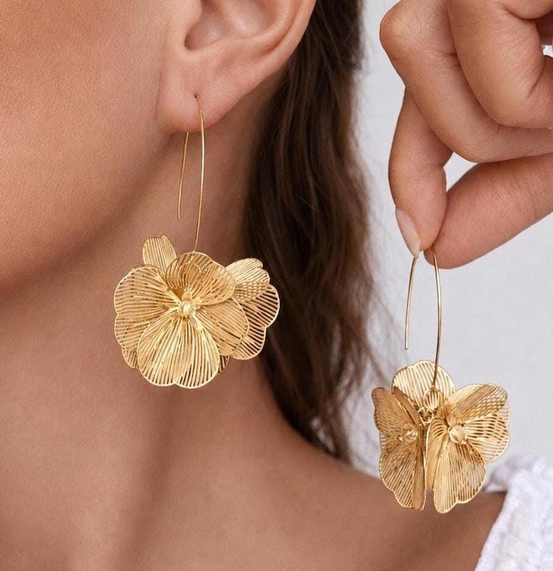 K&ouml;nnte beinhalten: Goldfarbene Blumenohrringe. Die Ohrringe haben ein filigranes, durchbrochenes Design in Form einer Blume. Sie sind an einem d&uuml;nnen, gebogenen Draht aufgeh&auml;ngt. Die Ohrringe werden in einer Nahaufnahme gezeigt, die die komplizierten Details des Designs hervorhebt.