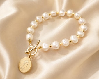 May include: A bracelet featuring white freshwater pearls interspersed with small gold-toned metal beads. The piece includes a gold-toned toggle clasp and a circular gold-toned pendant embossed with a profile portrait of a monarch.