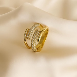 May include: A gold-colored ring with a unique design. The ring features two curved bands, one of which is encrusted with small, sparkling stones. The ring is set against a soft, cream-colored background.