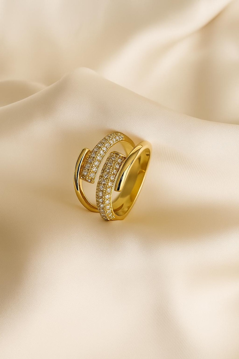 May include: A gold-colored ring with a unique design. The ring features two curved bands, one of which is encrusted with small, sparkling stones. The ring is set against a soft, cream-colored background.