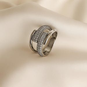 May include: A silver ring with a unique design. The ring features two bands, one of which is encrusted with small, sparkling stones. The ring is set against a neutral background, highlighting its elegant and modern aesthetic. A piece of jewelry.