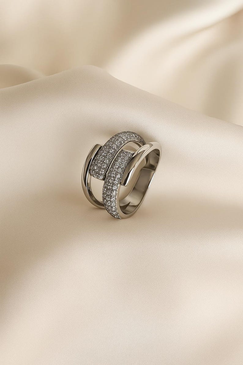 May include: A silver ring with a unique design. The ring features two bands, one of which is encrusted with small, sparkling stones. The ring is set against a neutral background, highlighting its elegant and modern aesthetic. A piece of jewelry.