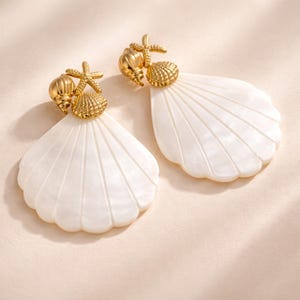 May include: A pair of gold-colored seashell earrings with white, fan-shaped pendants. Each earring features a small gold starfish, a seashell, and a conch shell. The earrings are designed to resemble a beach theme.