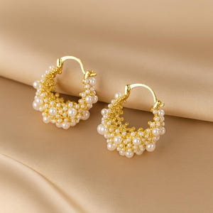 May include: A pair of gold hoop earrings featuring a textured, branch-like metal base densely covered in small, round white pearls. The earrings have a hinged closure mechanism and a circular shape, showcasing a cluster of pearls.