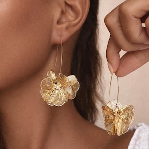 K&ouml;nnte beinhalten: Goldfarbene Blumenohrringe mit filigranem, offenem Design. Ein Ohrring wird getragen, der andere wird von einer Hand gehalten. Die Ohrringe haben einen langen, d&uuml;nnen Haken und eine dreidimensionale Blumenform.