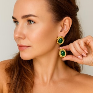 May include: Gold-toned, oval-shaped earrings with a vibrant green center stone. The earrings have a textured, twisted rope design around the gemstone. One earring is worn, and the other is held up to the ear.