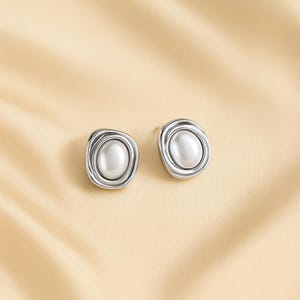 May include: A pair of silver-toned stud earrings. Each earring features a central, oval-shaped, white pearl surrounded by a textured, layered silver frame. The earrings are displayed on a light beige background.