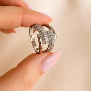 May include: A silver ring with a geometric design. The ring features a central rectangular section encrusted with small, sparkling stones, flanked by smooth, curved bands. The ring is held between two fingers against a neutral background.