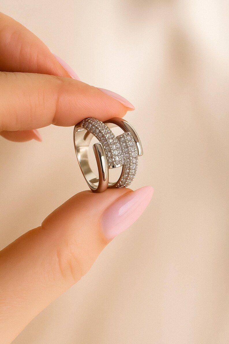 May include: A silver ring with a geometric design. The ring features a central rectangular section encrusted with small, sparkling stones, flanked by smooth, curved bands. The ring is held between two fingers against a neutral background.