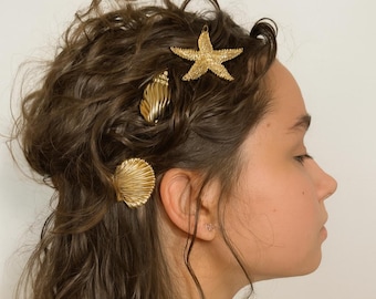 May include: Three gold-toned hair clips shaped like marine life, including a textured starfish, a spiral conch shell, and a ridged scallop shell, are secured in wavy brown hair, adding metallic accents to the hairstyle.