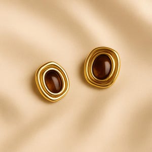 May include: A pair of gold-toned, oval-shaped clip-on earrings. Each earring features a brown, oval-shaped stone set within a layered gold frame. The earrings have a vintage aesthetic.