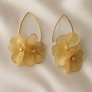 K&ouml;nnte beinhalten: Ein Paar goldfarbene Blumenohrringe. Jeder Ohrring hat drei zarte, drahtartige Bl&uuml;tenbl&auml;tter mit einem kleinen, funkelnden Edelstein in der Mitte. Die Ohrringe h&auml;ngen an einem goldfarbenen, tropfenf&ouml;rmigen Haken. Der Hintergrund ist ein weicher, cremefarbener Stoff.