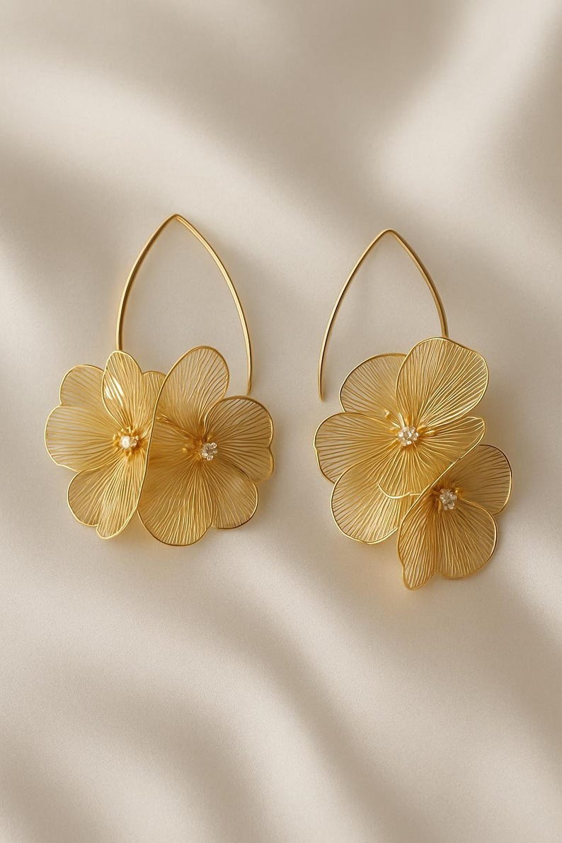 K&ouml;nnte beinhalten: Ein Paar goldfarbene Blumenohrringe. Jeder Ohrring hat drei zarte, drahtartige Bl&uuml;tenbl&auml;tter mit einem kleinen, funkelnden Edelstein in der Mitte. Die Ohrringe h&auml;ngen an einem goldfarbenen, tropfenf&ouml;rmigen Haken. Der Hintergrund ist ein weicher, cremefarbener Stoff.