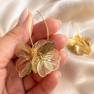 K&ouml;nnte beinhalten: Goldfarbene Blumen-Creolen. Die Ohrringe haben ein zartes, mehrschichtiges Blumendesign mit filigranen Details. Die Ohrringe werden in einer Hand vor einem weichen, cremefarbenen Hintergrund gehalten. Die Ohrringe sind ein stilvolles Accessoire.