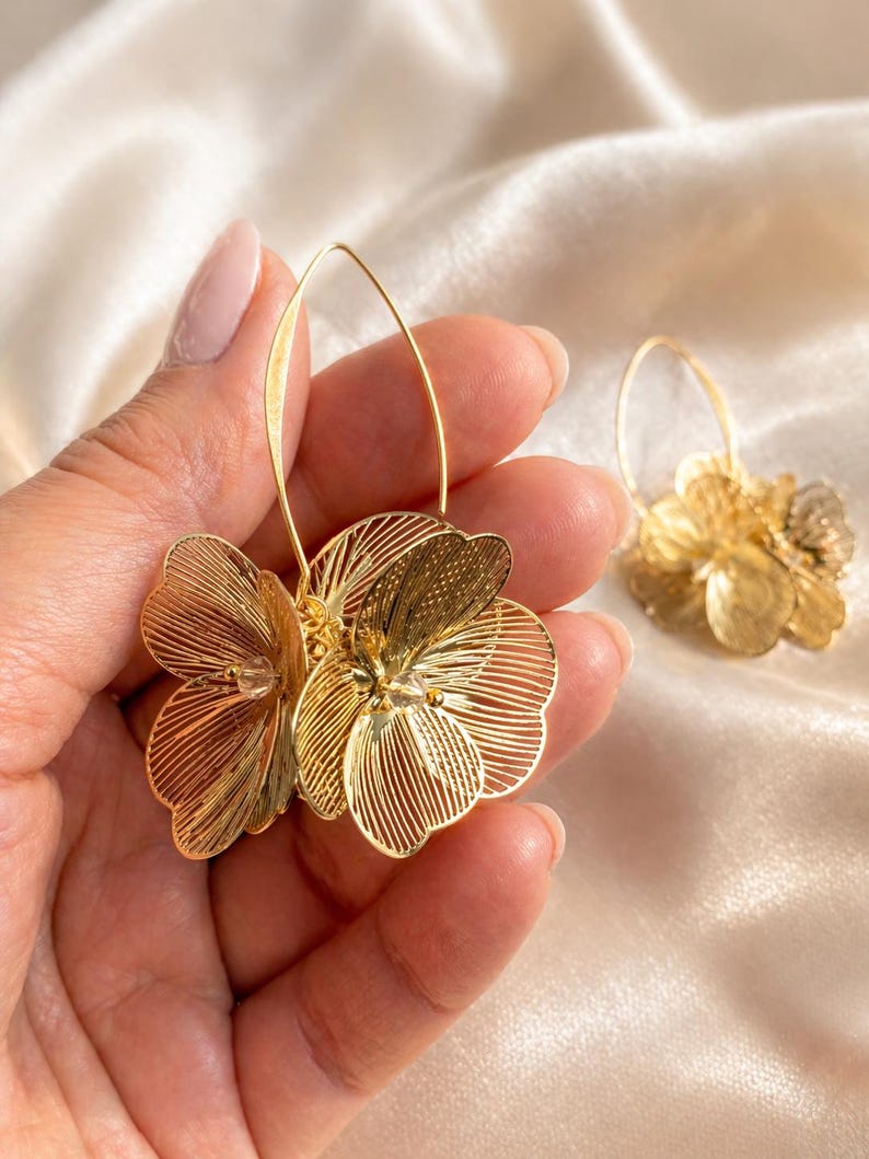 K&ouml;nnte beinhalten: Goldfarbene Blumen-Creolen. Die Ohrringe haben ein zartes, mehrschichtiges Blumendesign mit filigranen Details. Die Ohrringe werden in einer Hand vor einem weichen, cremefarbenen Hintergrund gehalten. Die Ohrringe sind ein stilvolles Accessoire.