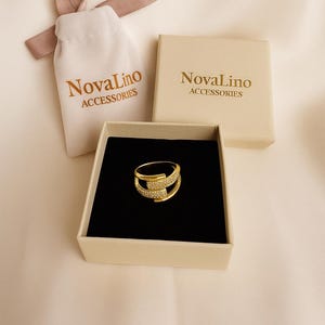 May include: A gold-colored ring with a unique design, set in a black velvet-lined box. The ring is adorned with small, sparkling stones. The image also shows a small drawstring bag and a box, both with the text "NovaLino ACCESSORIES".
