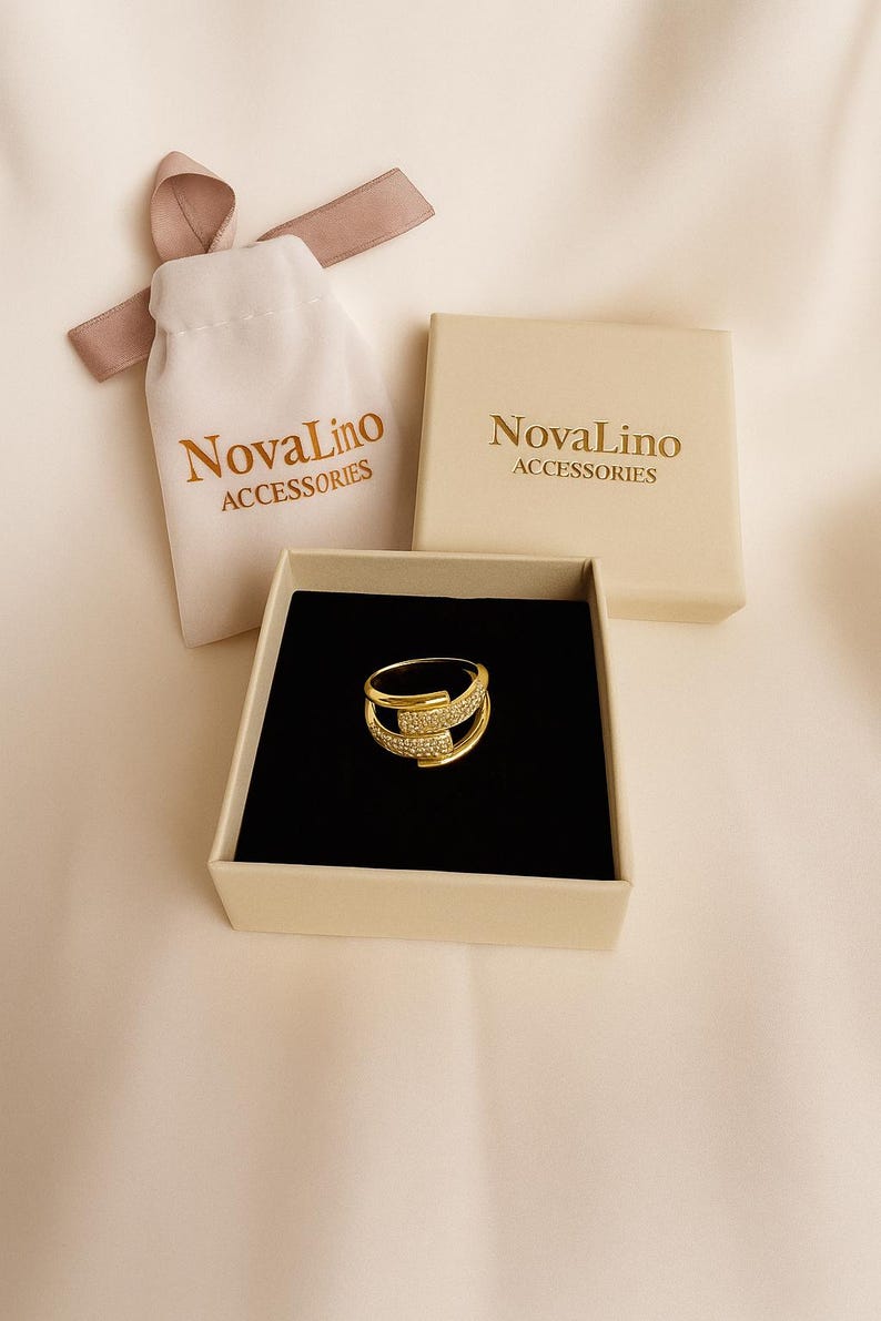 May include: A gold-colored ring with a unique design, set in a black velvet-lined box. The ring is adorned with small, sparkling stones. The image also shows a small drawstring bag and a box, both with the text "NovaLino ACCESSORIES".