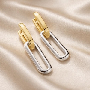 May include: A pair of gold and silver-toned earrings. The top portion of the earrings is gold, while the elongated oval drop is silver. The earrings are displayed on a cream-colored, satin-like fabric.
