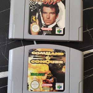 Może przedstawiać: Dwie szare kasety z grami wideo na konsolę Nintendo 64. Górna kaseta posiada etykietę gry GoldenEye 007 z wizerunkiem mężczyzny trzymającego pistolet. Dolna kaseta posiada etykietę gry Command & Conquer z grafiką przedstawiającą wybuch.