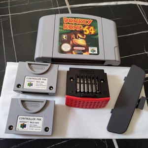 Peut inclure: Une cartouche de jeu vidéo Donkey Kong 64 grise pour Nintendo 64, deux cartes mémoire Controller Pak grises, un expansion pak noir et rouge, et un couvercle en plastique gris. Tous les articles sont des accessoires originaux Nintendo 64.