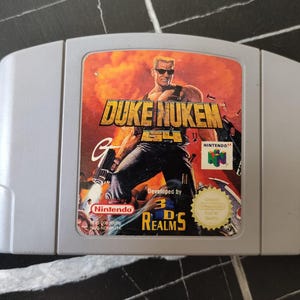 Könnte beinhalten: Eine graue Nintendo 64 Videospielkassette für Duke Nukem 64. Das Etikett zeigt eine zentrale Figur mit Sonnenbrille vor einem orangefarbenen Explosionshintergrund sowie den Text Duke Nukem 64, Nintendo, 3D Realms und ein goldenes Qualitätssiegel.