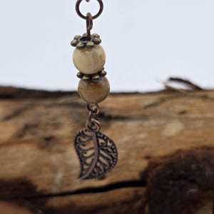 Può includere: Un ciondolo fatto a mano con due perline rotonde di colore chiaro separate da un distanziatore decorativo e un ciondolo a forma di foglia color rame dettagliato. Il ciondolo è appeso a un anello color rame.