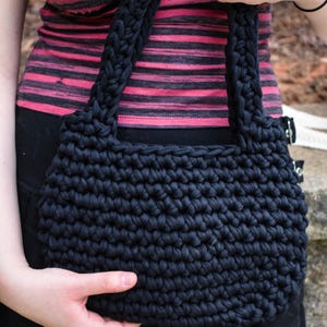 May include: A black, hand-crocheted tote bag with thick, braided handles. The bag has a textured, chunky stitch pattern. The person holding the bag is wearing a pink and black striped top.