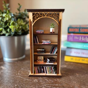 May include: A miniature wooden bookshelf with several shelves, filled with tiny books and a small potted plant. The bookshelf has a dark brown top and decorative cutouts. A stack of full-sized books is visible in the background.