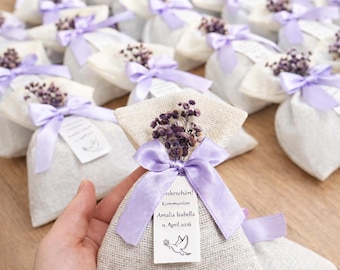 May include: A small beige burlap pouch tied with a light purple satin ribbon and decorated with a sprig of dried purple flowers. A white rectangular tag is attached, featuring the text "Dankeschön! Kommunion Amalia Isabella 11. April 2026" and a dove.