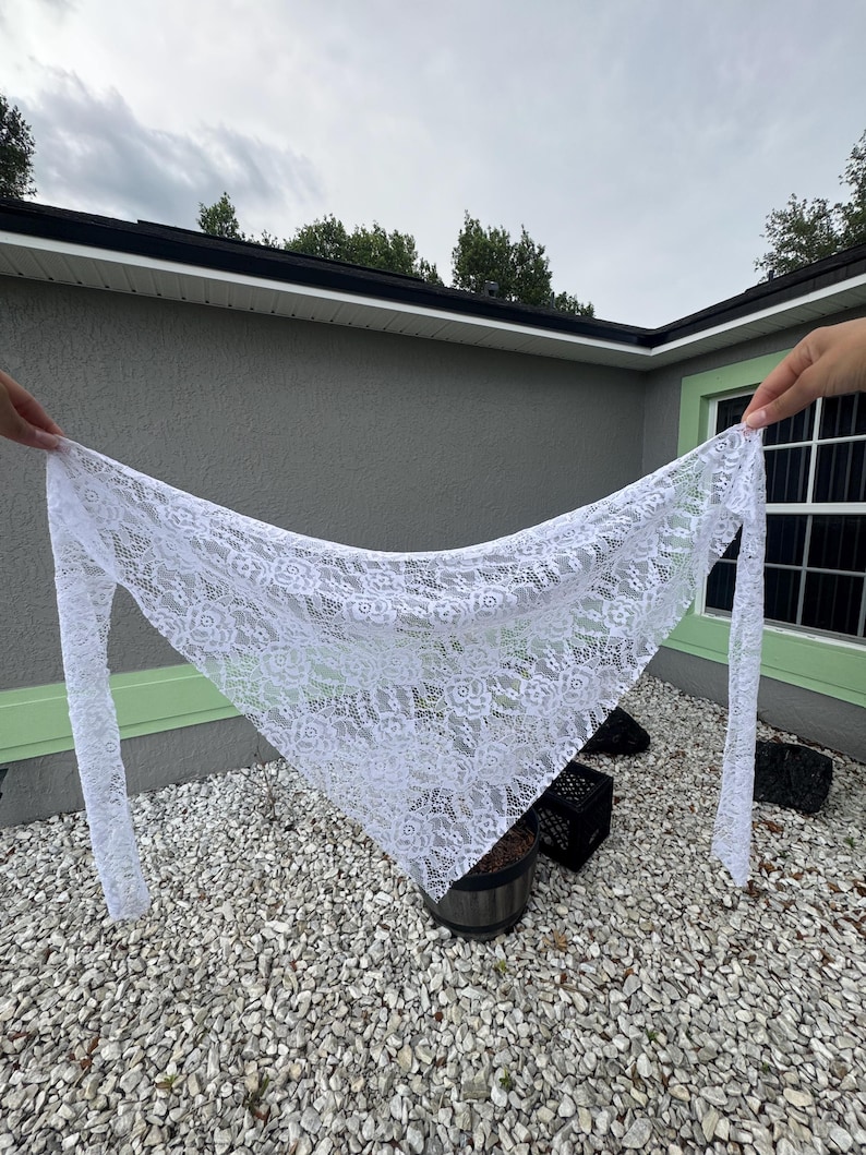 Puede incluir: Un chal de encaje blanco con un estampado floral se sostiene contra un cielo nublado. La bufanda triangular tiene mangas largas y estrechas y est&aacute; hecha de una tela delicada y transparente. El chal se exhibe al aire libre.