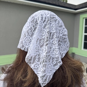 Puede incluir: Un velo blanco de encaje, con un dise&ntilde;o floral, est&aacute; drapeado sobre la cabeza. La delicada tela es visible contra el fondo del cabello casta&ntilde;o. El velo tiene forma cuadrada.
