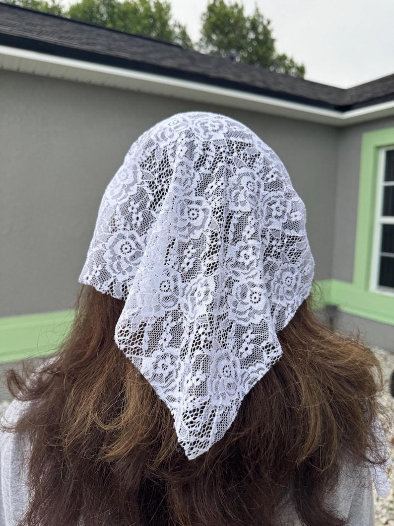 Puede incluir: Un velo blanco de encaje, con un dise&ntilde;o floral, est&aacute; drapeado sobre la cabeza. La delicada tela es visible contra el fondo del cabello casta&ntilde;o. El velo tiene forma cuadrada.
