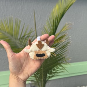 Könnte beinhalten: Ein Appa-Ornament aus Filz mit cremefarbenem Körper, braunen Hörnern und Pfeil sowie einem schwarzen Mund. Das Ornament hat eine weiße Schlaufe zum Aufhängen. Im Hintergrund sind Palmenwedel und eine graue Wand zu sehen.