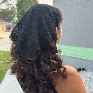 May include: A black triangular headscarf made of delicate lace fabric with a floral pattern. The headscarf is tied at the back of the head, covering the top portion of the hair, and features scalloped edges along the perimeter of the fabric.